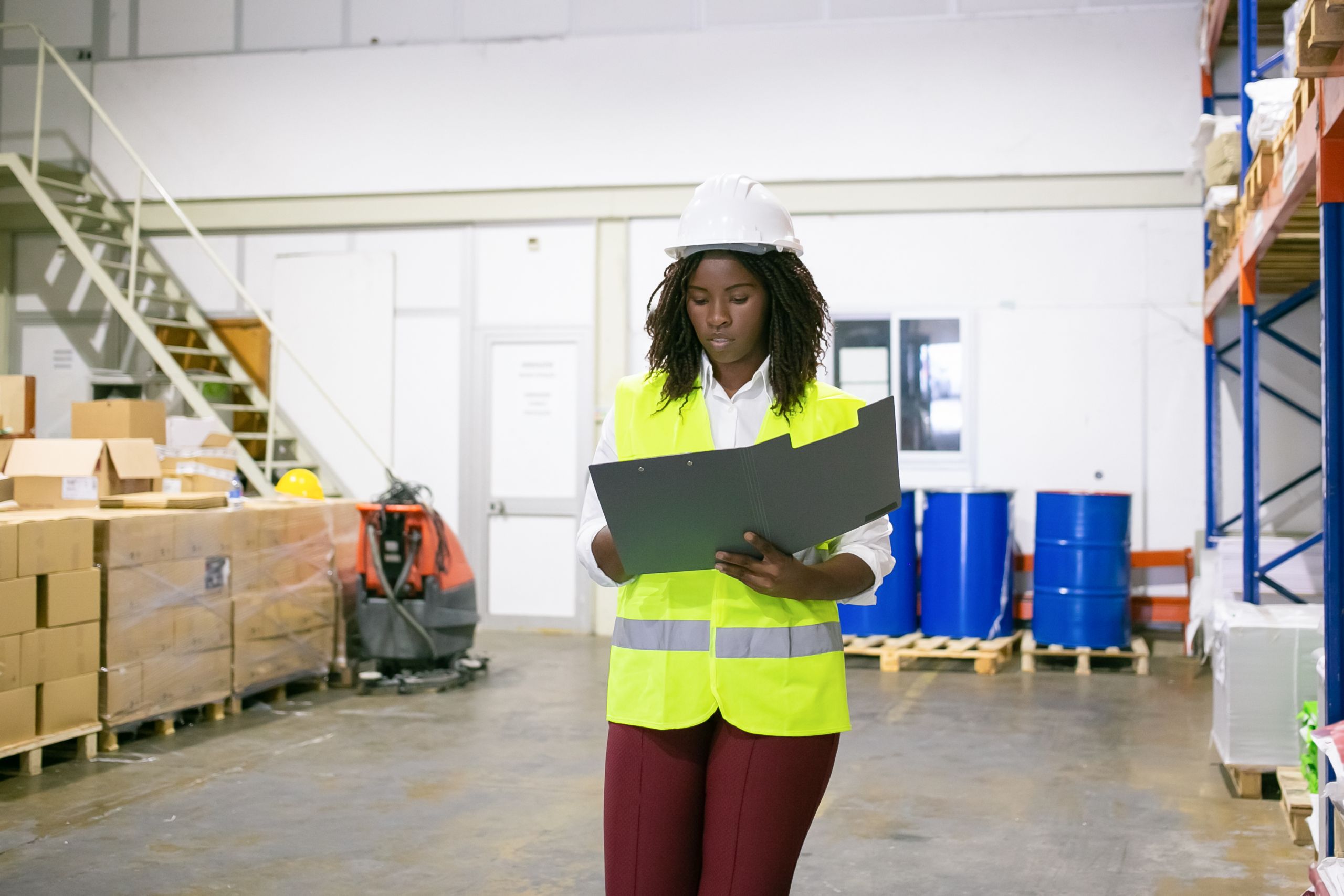 Focused female logistic employee in hardhat and safety vest walking in warehouse, carrying open folder, looking through document. Copy space, front view. Labor and inspection concept